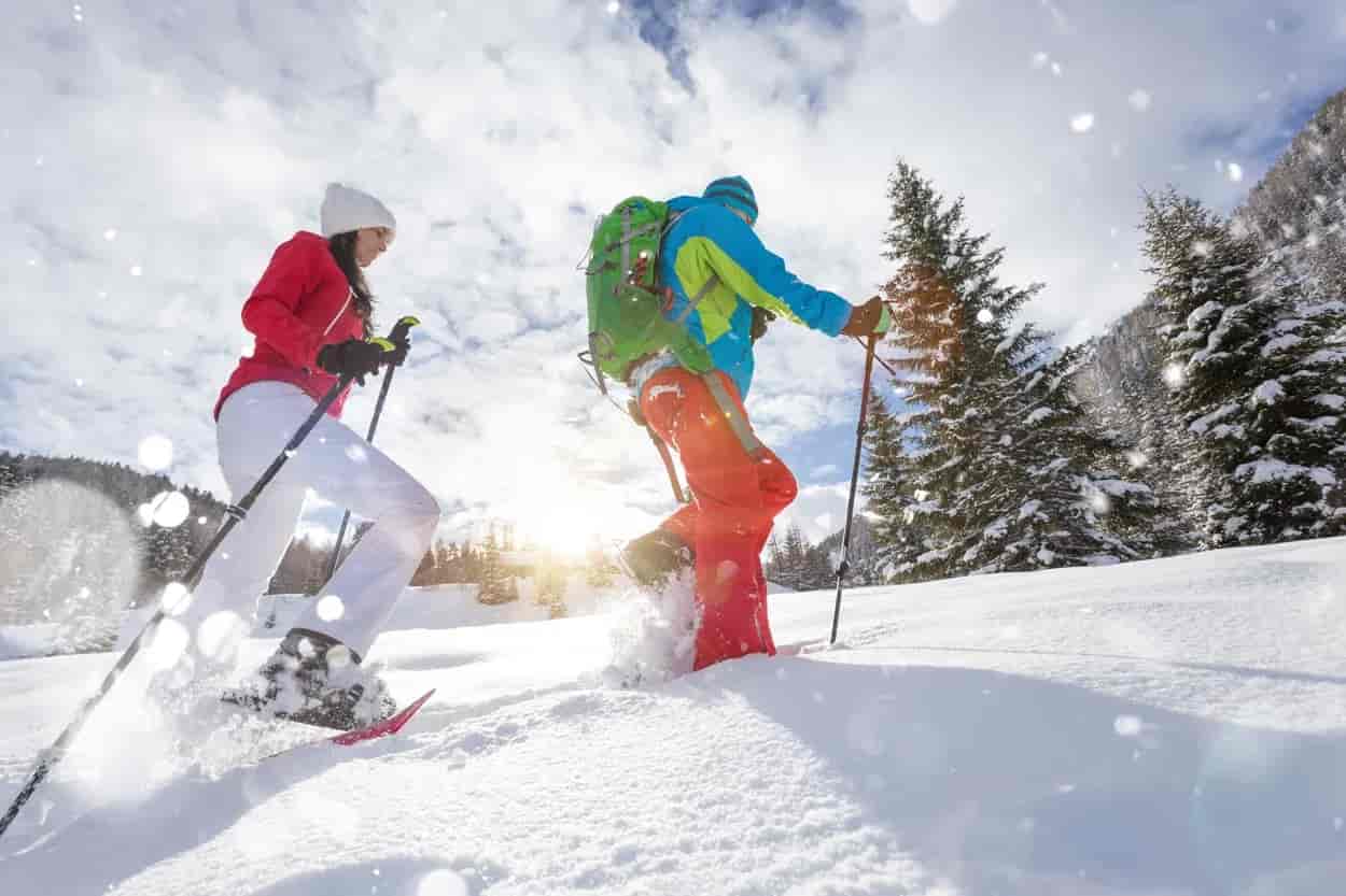 Two people snowshoeing uphill through deep snow with evergreen trees and bright sun.