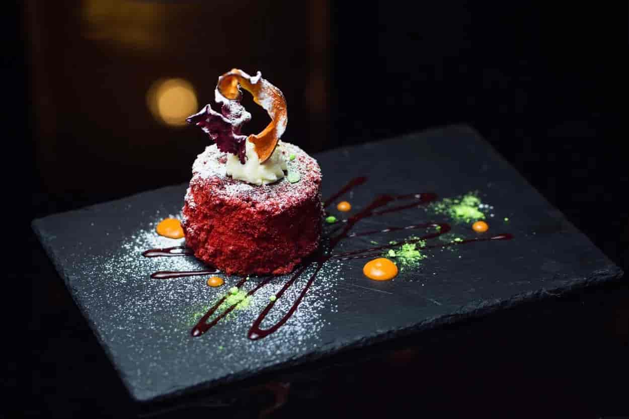 Red velvet dessert with cream and garnish served on a dark slate plate with sauces.