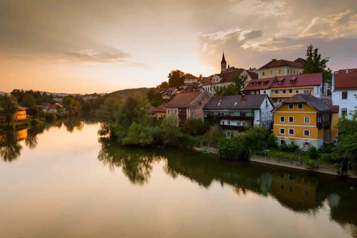 Town houses along a river reflecting golden sunset light, with a church spire visible.