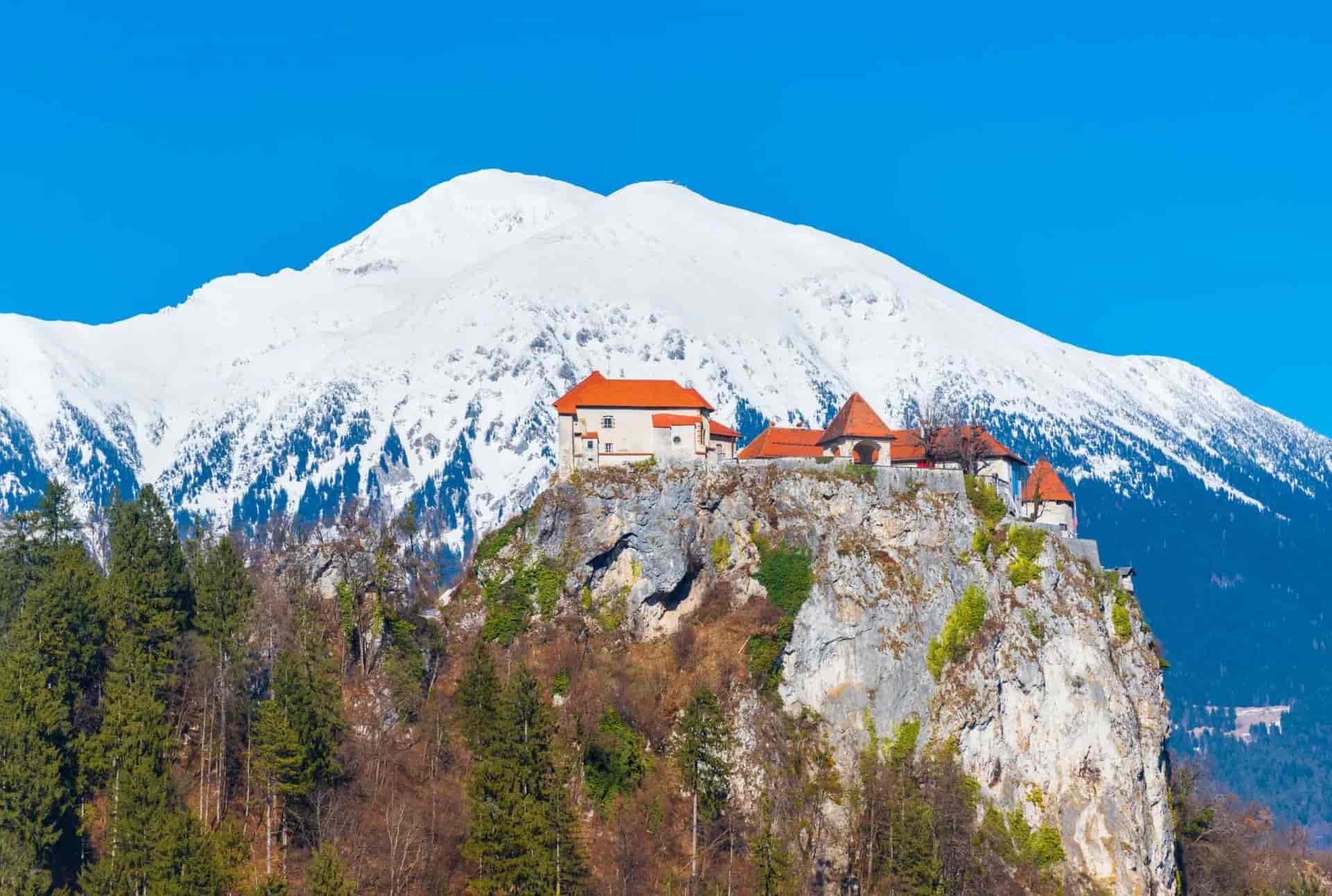 Bled Castle perched on cliff with snow-covered mountain and clear blue sky in winter