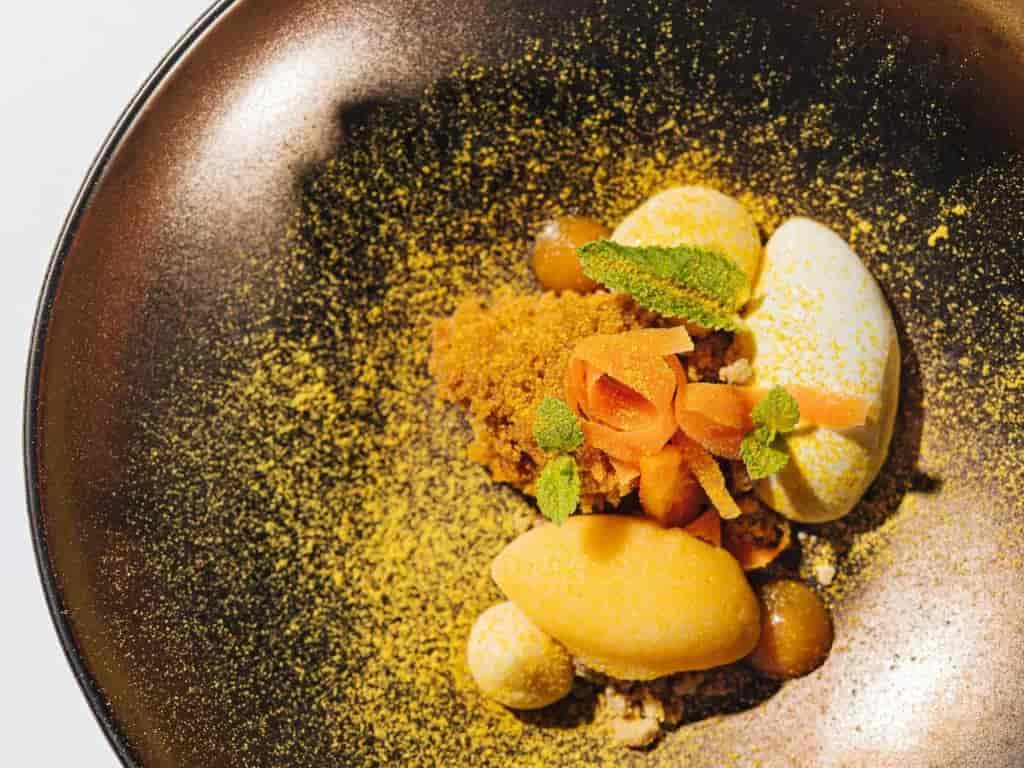 Gourmet dessert with yellow powder, quenelles, mint, and candied fruit on a dark plate.