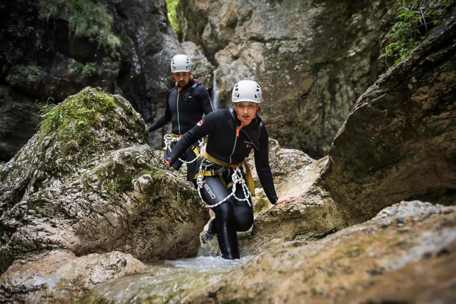 Canyoneering participants in wetsuits and helmets hiking through a rocky stream in a canyon.
