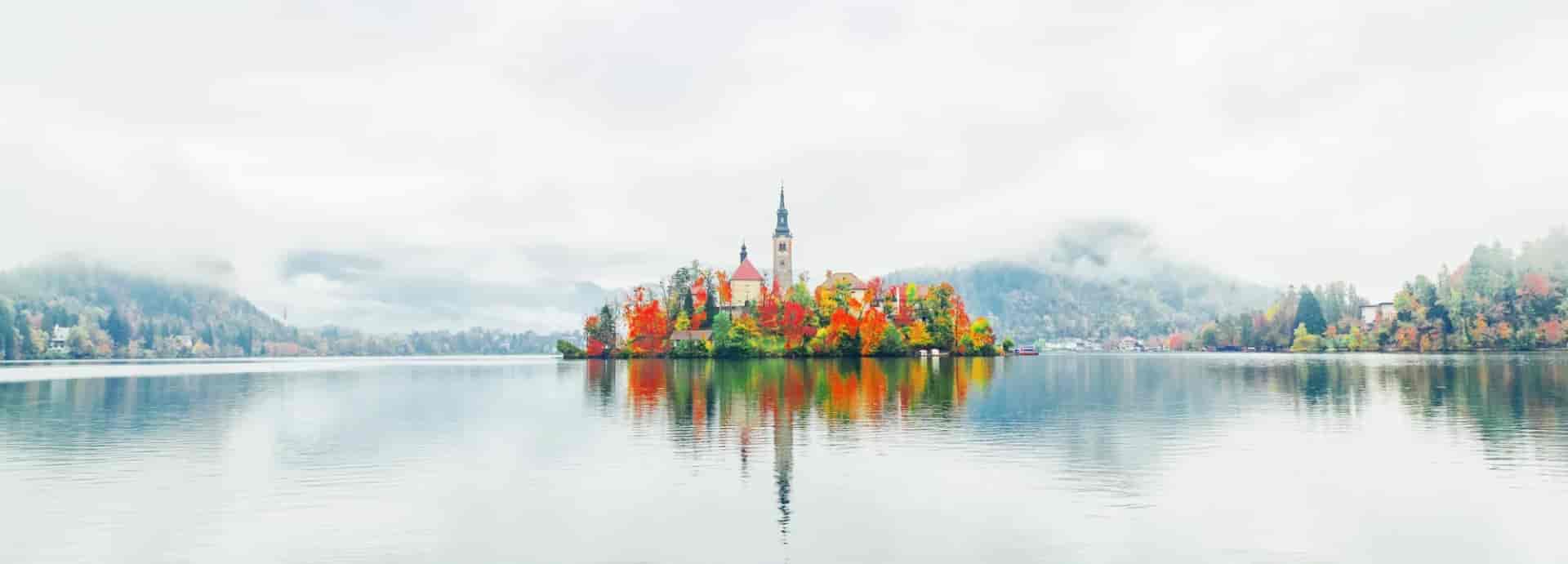 Lake Bled island church with autumn foliage reflected in calm water under misty mountains