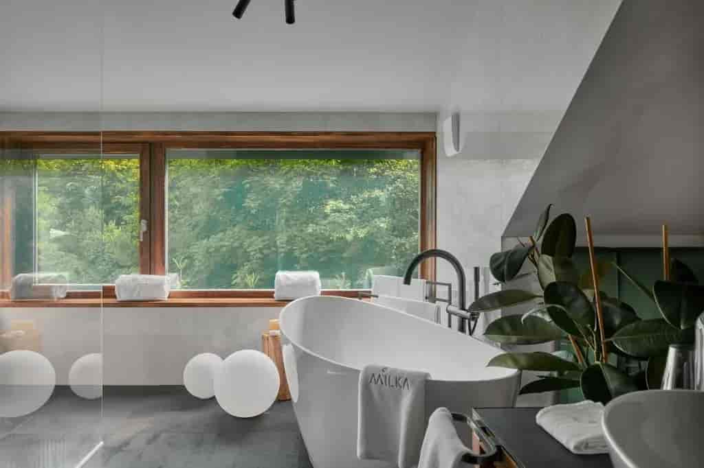 Modern bathroom with freestanding tub, large window overlooking lush green forest, and decorative orbs.