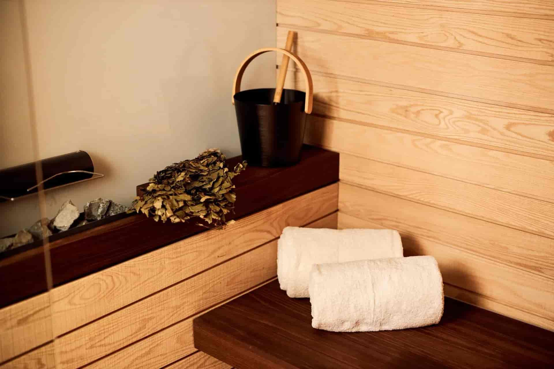 Sauna accessories: rolled white towels, black bucket, ladle, and birch whisk on wooden shelves.