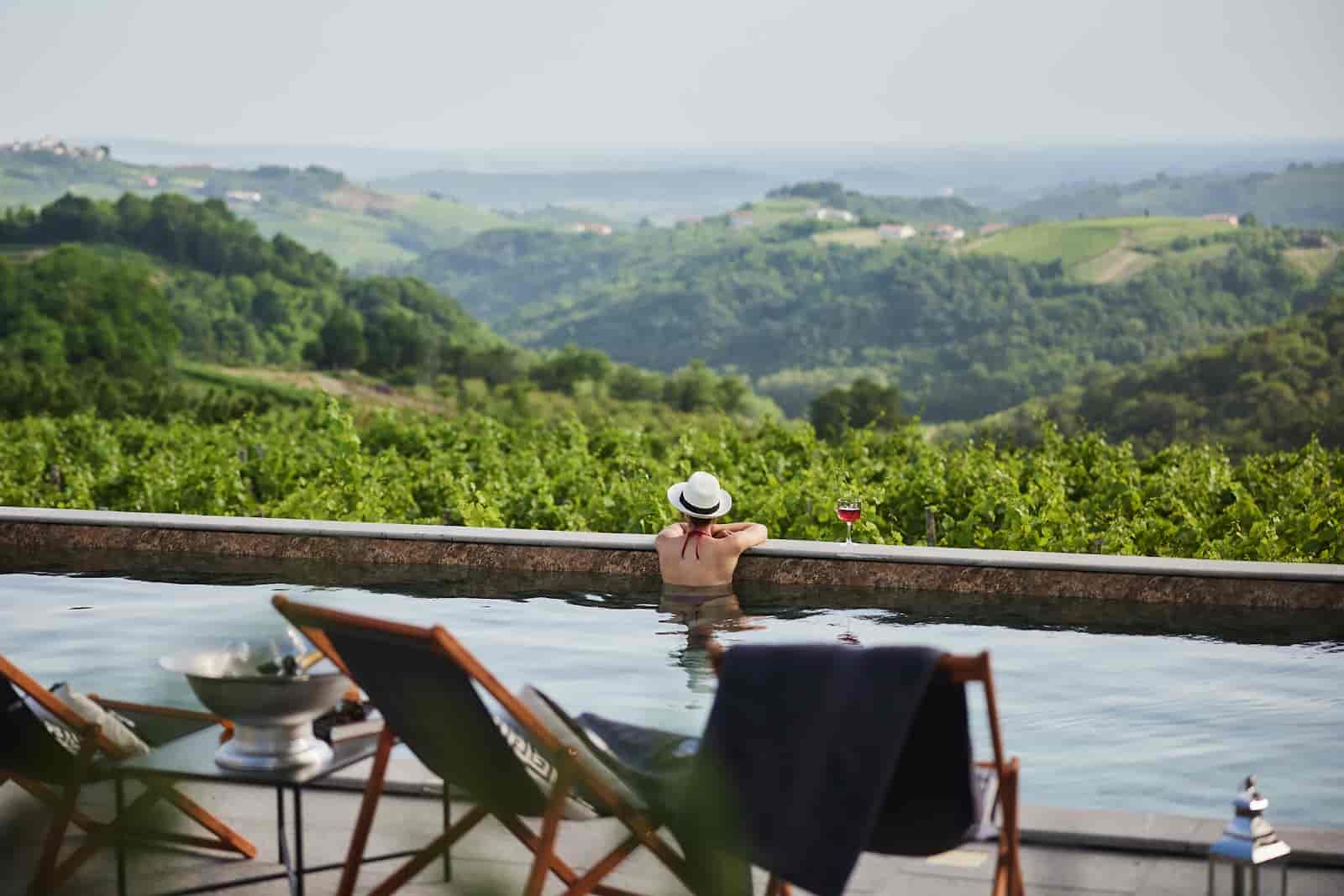Woman in infinity pool overlooking lush green vineyard hills with glass of wine.