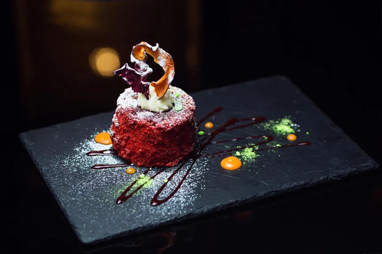 Red velvet cake dusted with powdered sugar on a black slate plate with sauce drizzles.