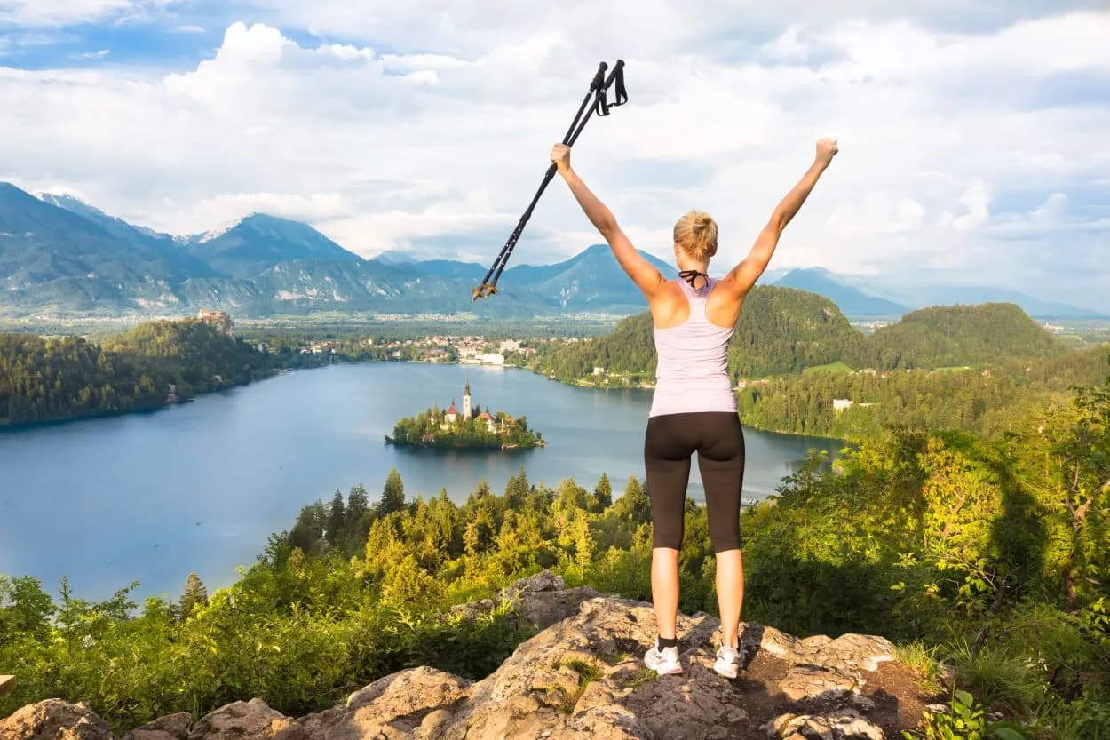 Hiker with trekking poles celebrating at viewpoint overlooking Lake Bled island and Julian Alps.