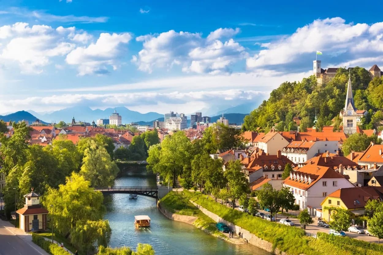 Ljubljana city center with riverboats, castle on hill, and mountains under blue sky.