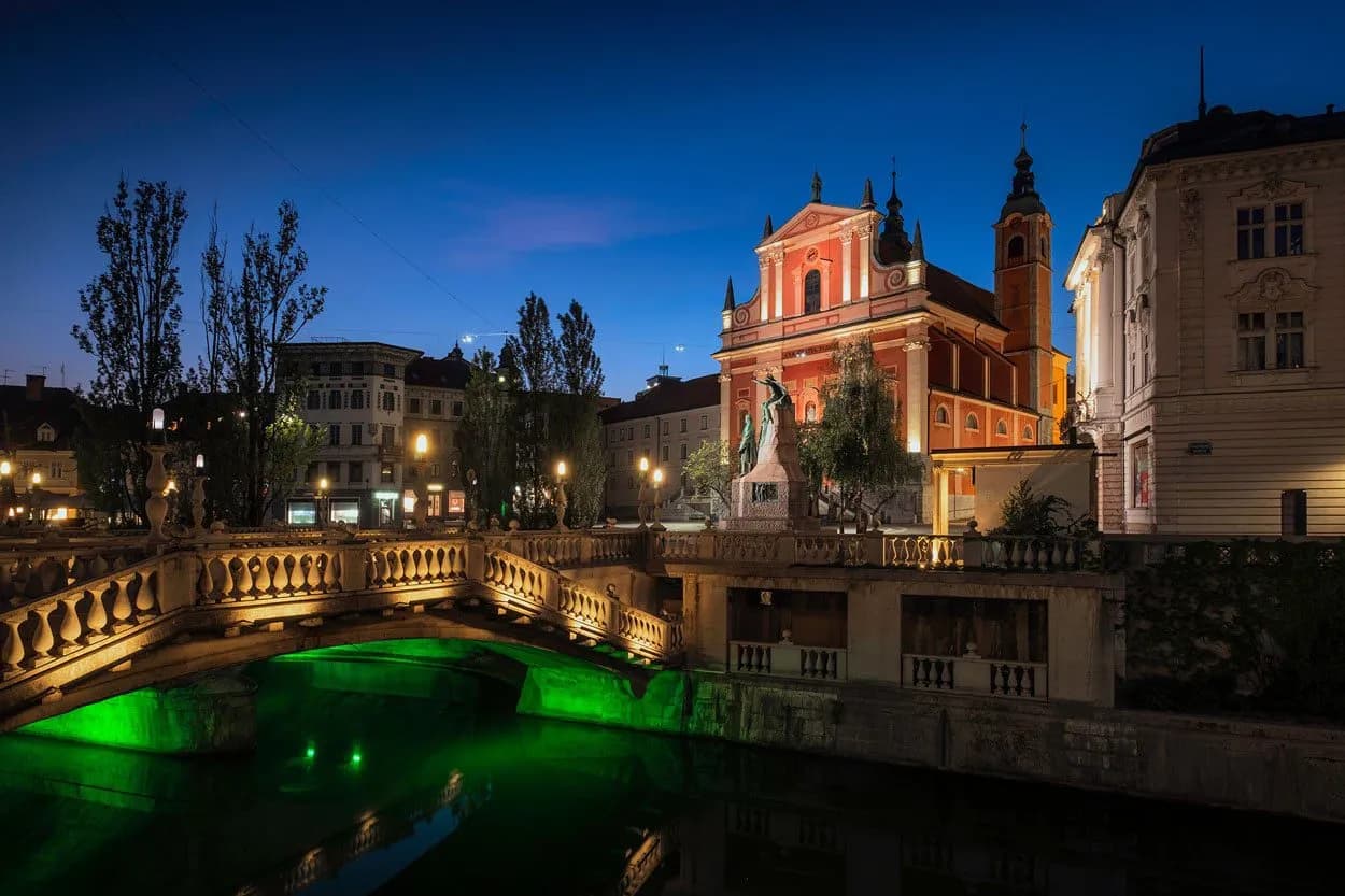 Triple Bridge and pink Franciscan Church illuminated at night in Ljubljana's Preseren Square.