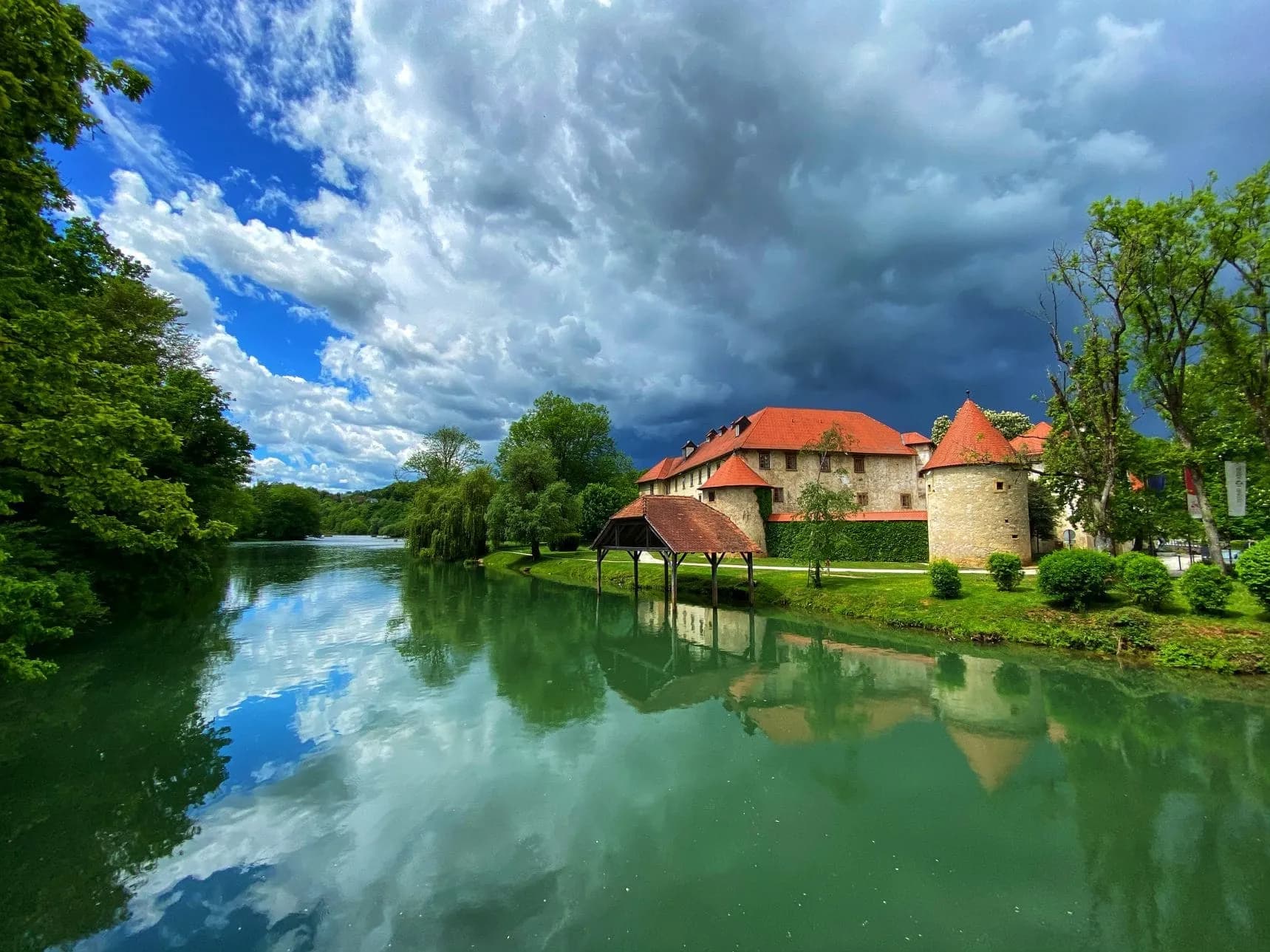 Otočec Castle in Dolenjska Region reflected in green river under dramatic cloudy sky.