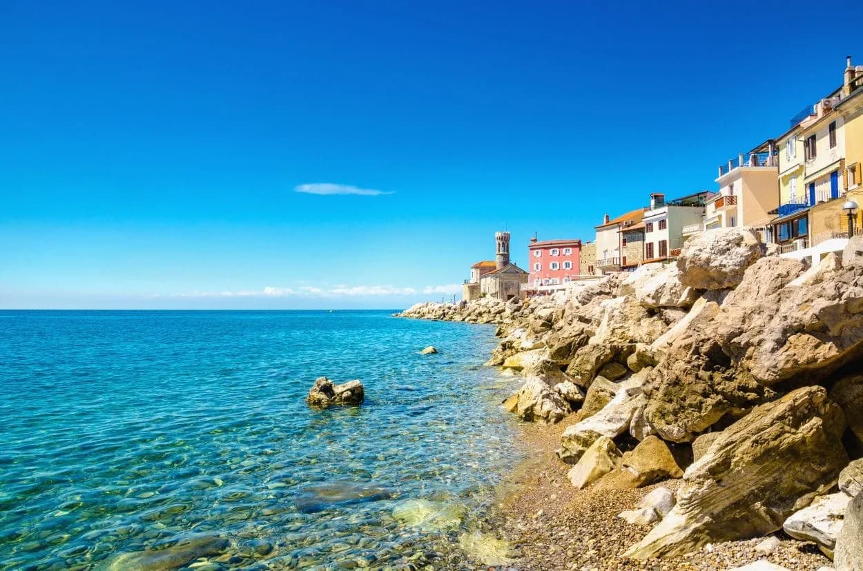 Rocky coastline meets clear turquoise sea below colorful waterfront buildings in Piran.