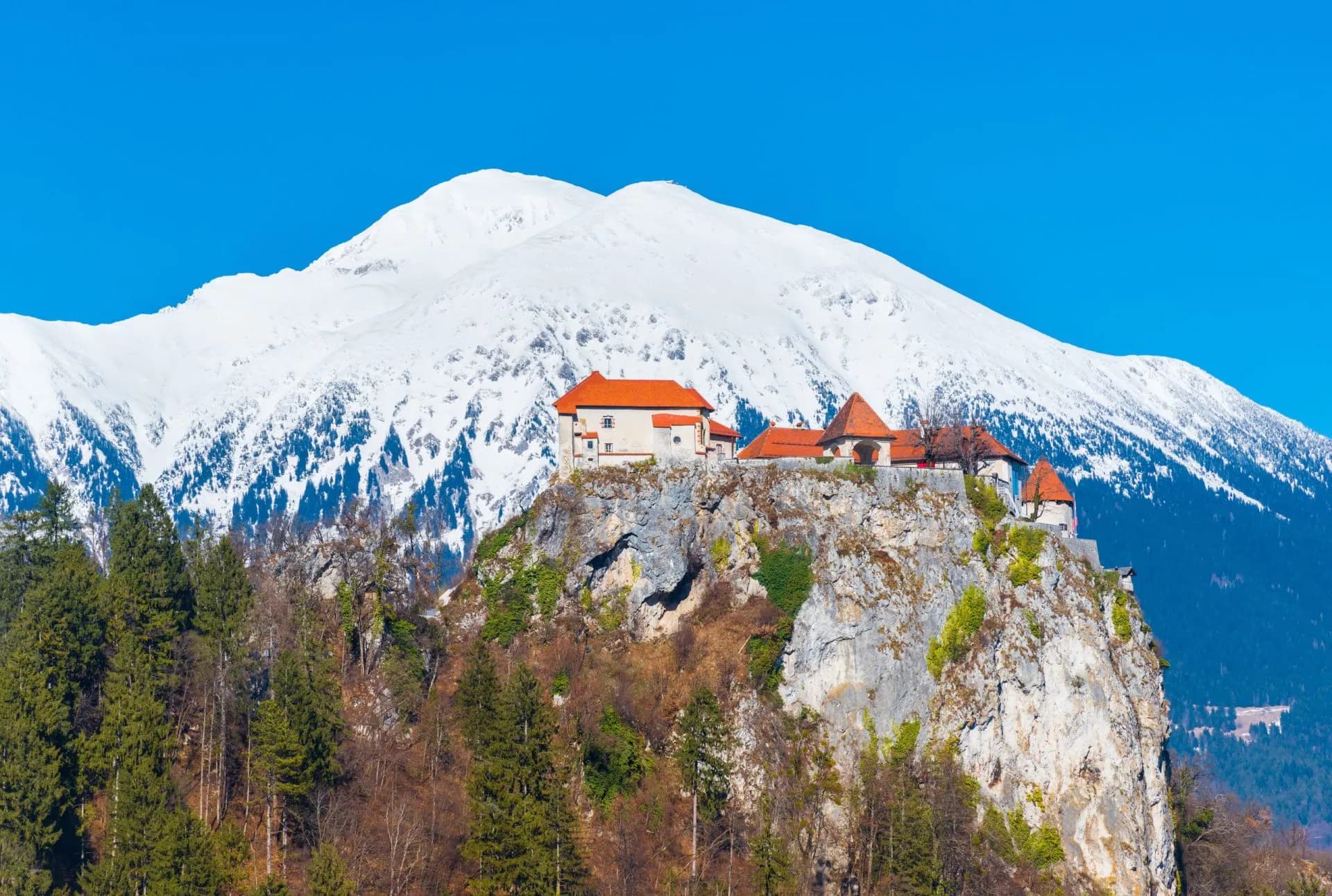 Bled Castle perched on cliff with snow-covered mountain and clear blue sky in winter