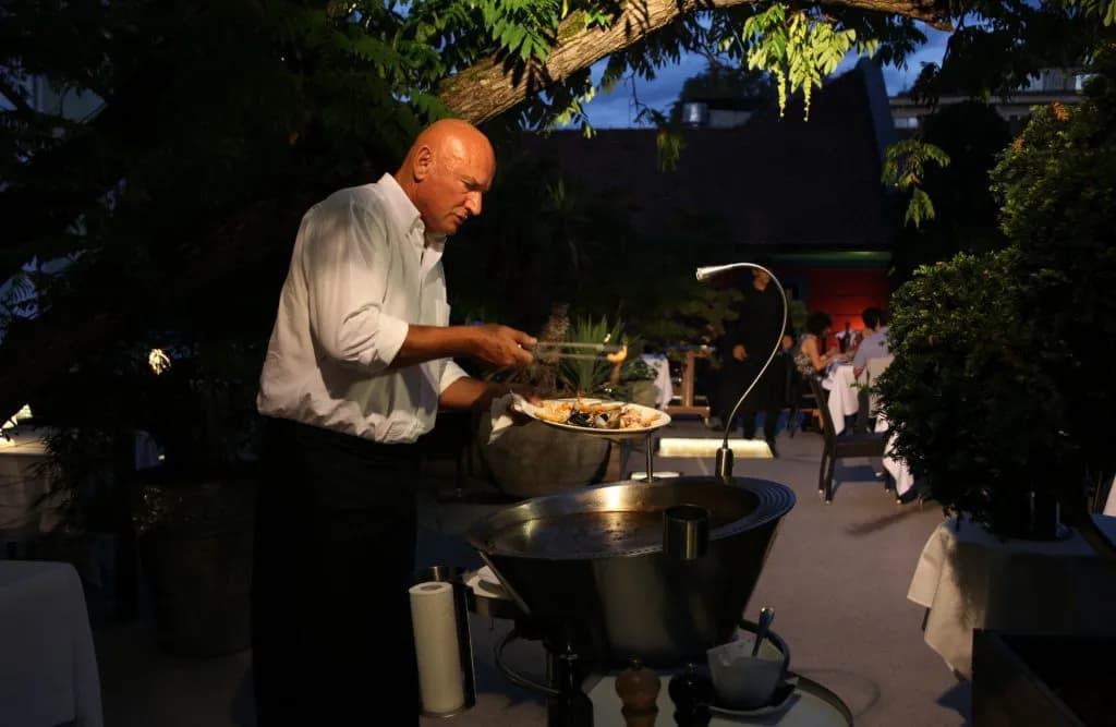 Chef serving seafood from a large pan at an outdoor evening dining area with foliage