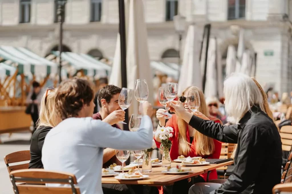 Group toasting with wine glasses at an outdoor cafe table with food in a sunny city square.