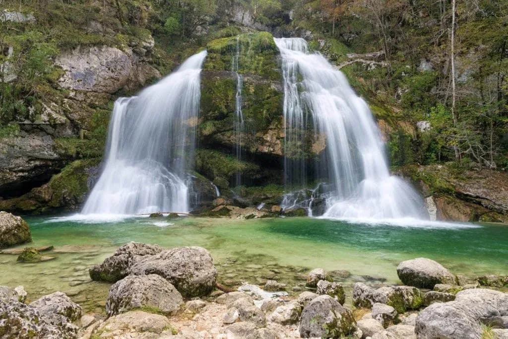 Wide waterfall cascading over mossy rocks into a clear, emerald pool with foreground boulders