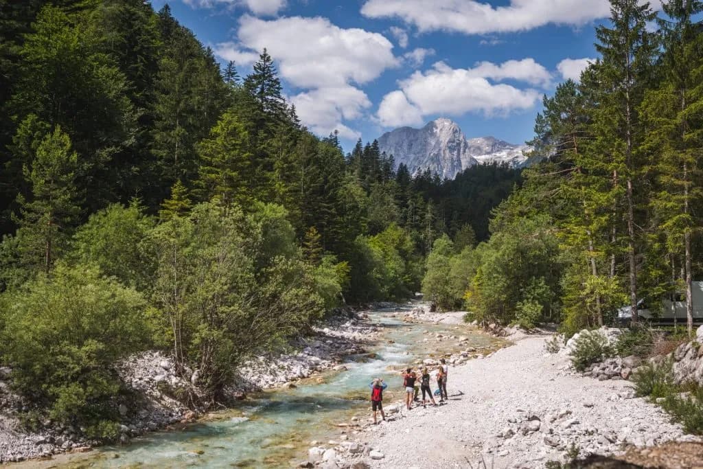 Hikers by clear river in Triglavska Bistrica valley with forested slopes and rocky mountain peaks.