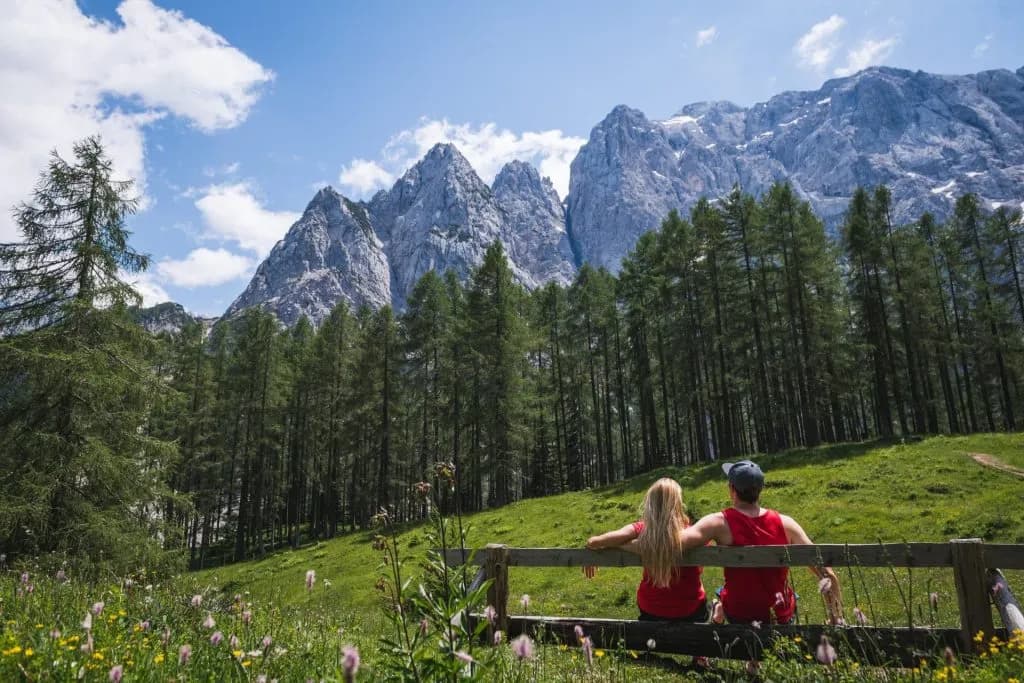 Couple sitting on a bench overlooking Vršič Pass mountains and pine forest in summer