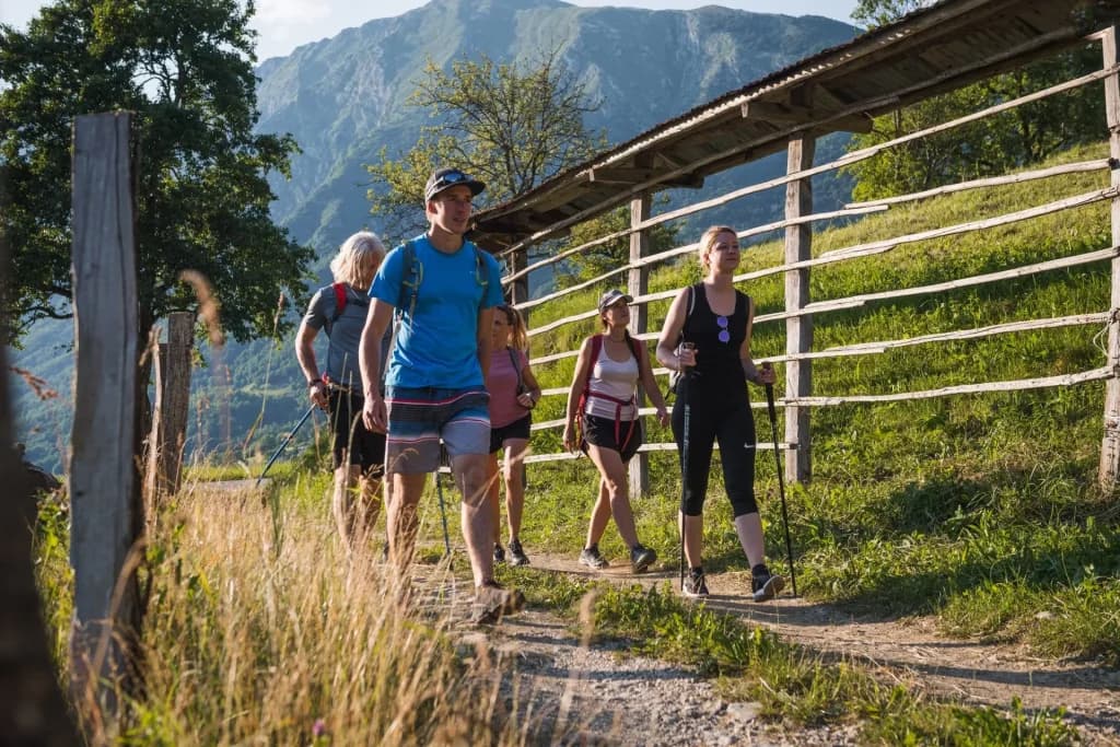 Hikers with poles walk on a dirt path near a wooden structure in Drežnica village with mountains in the background.