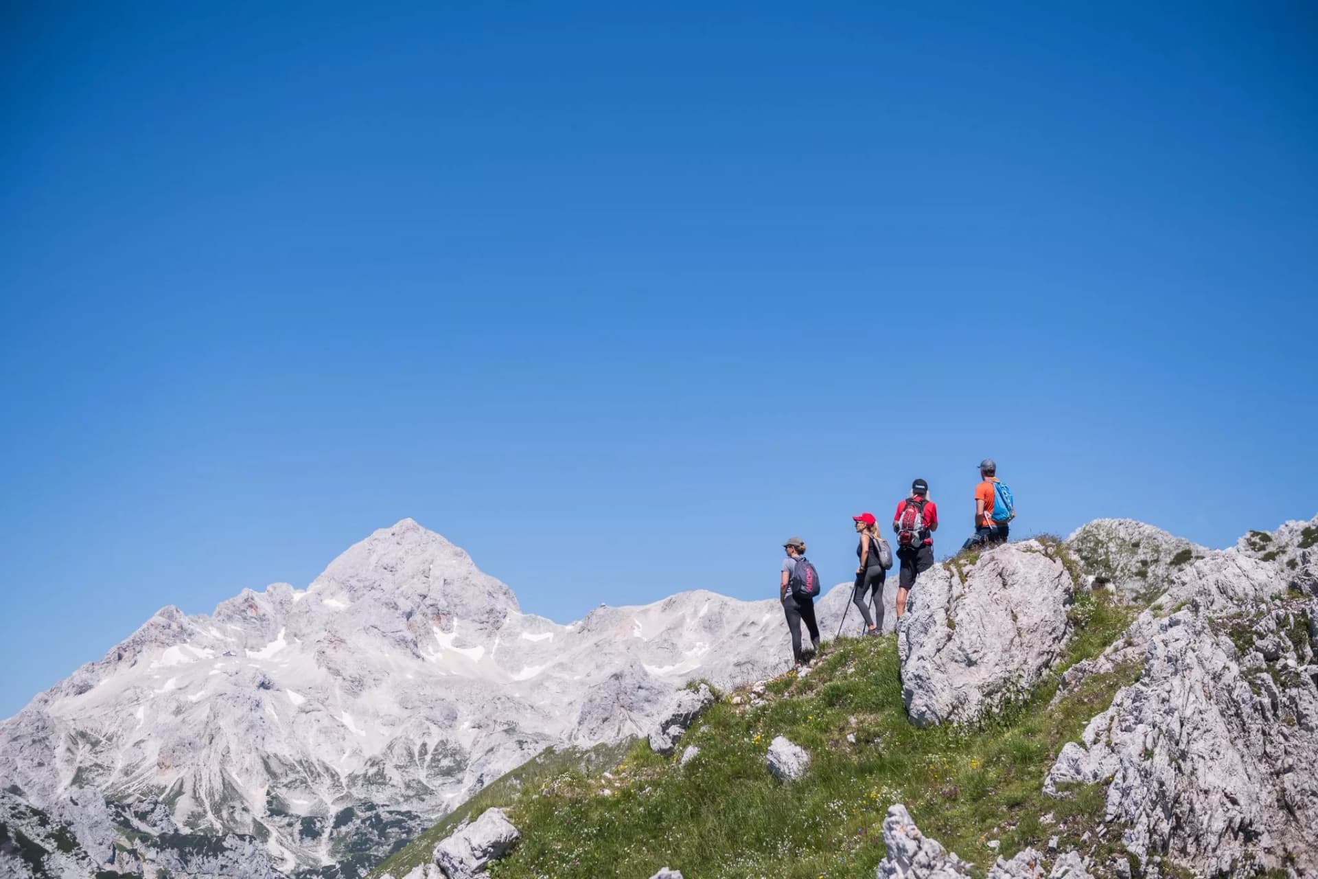 Hikers on grassy ridge overlooking snow-dusted mountains under clear blue sky in Slovenia.