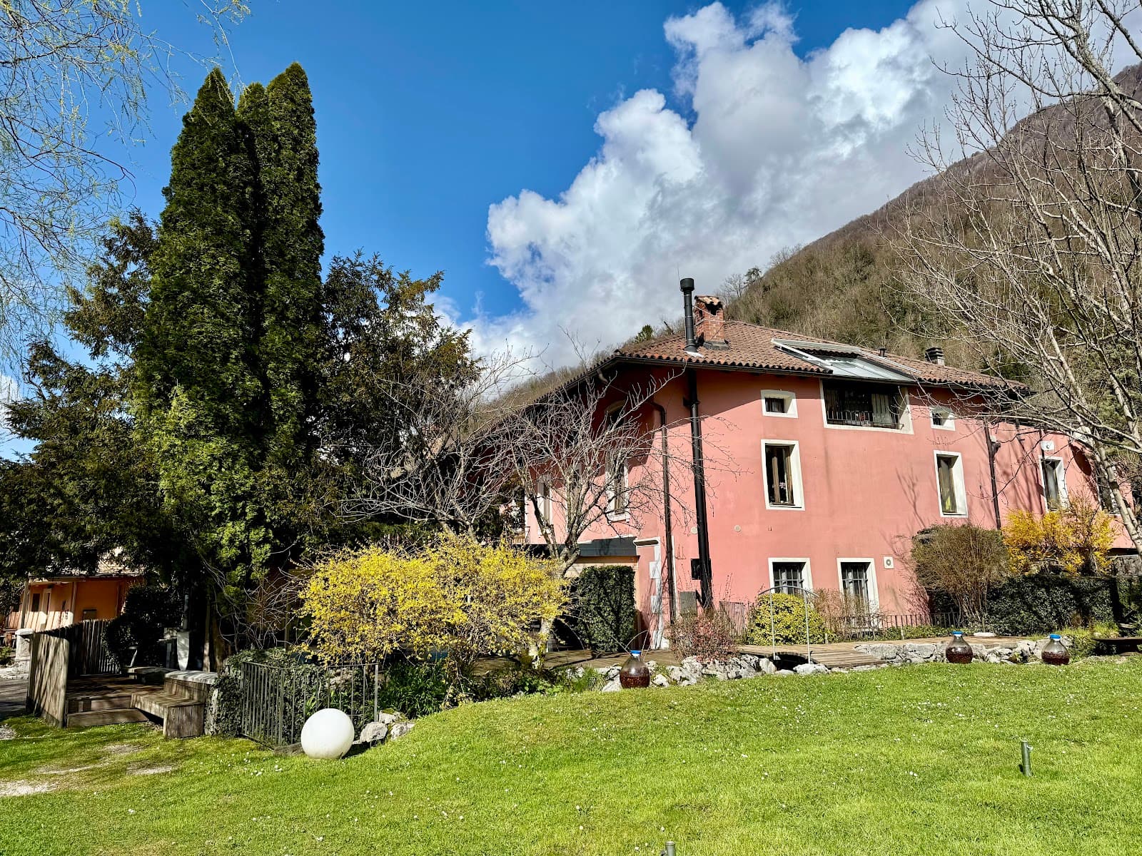 Pink house with garden, tall trees, and steep wooded hill under blue sky