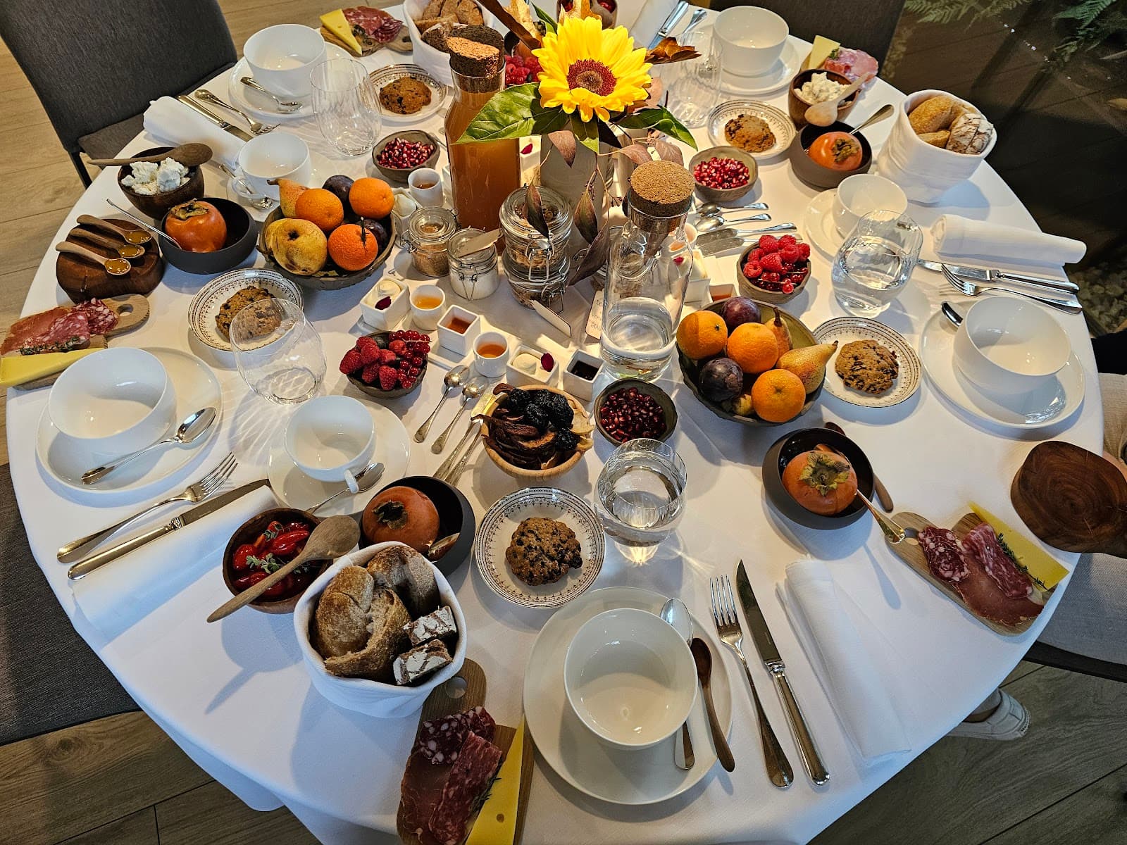 Lavish breakfast spread with fruit, bread, cheese, charcuterie, and a sunflower centerpiece on a white table.