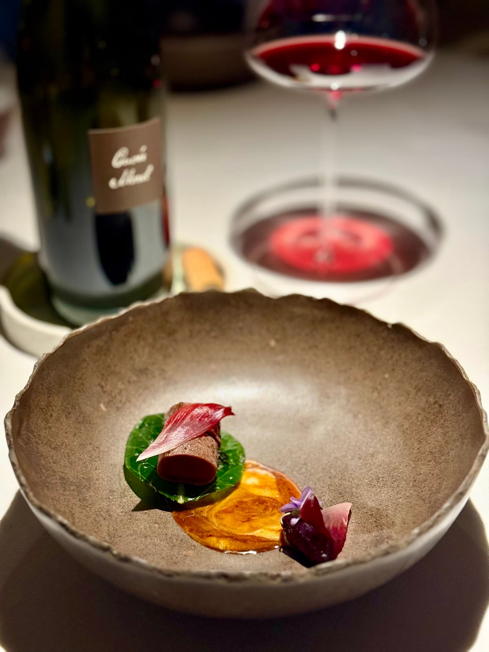 Gourmet dish with sauce, greens, and flower garnish, wine bottle, and glass in background at Hiša Franko.