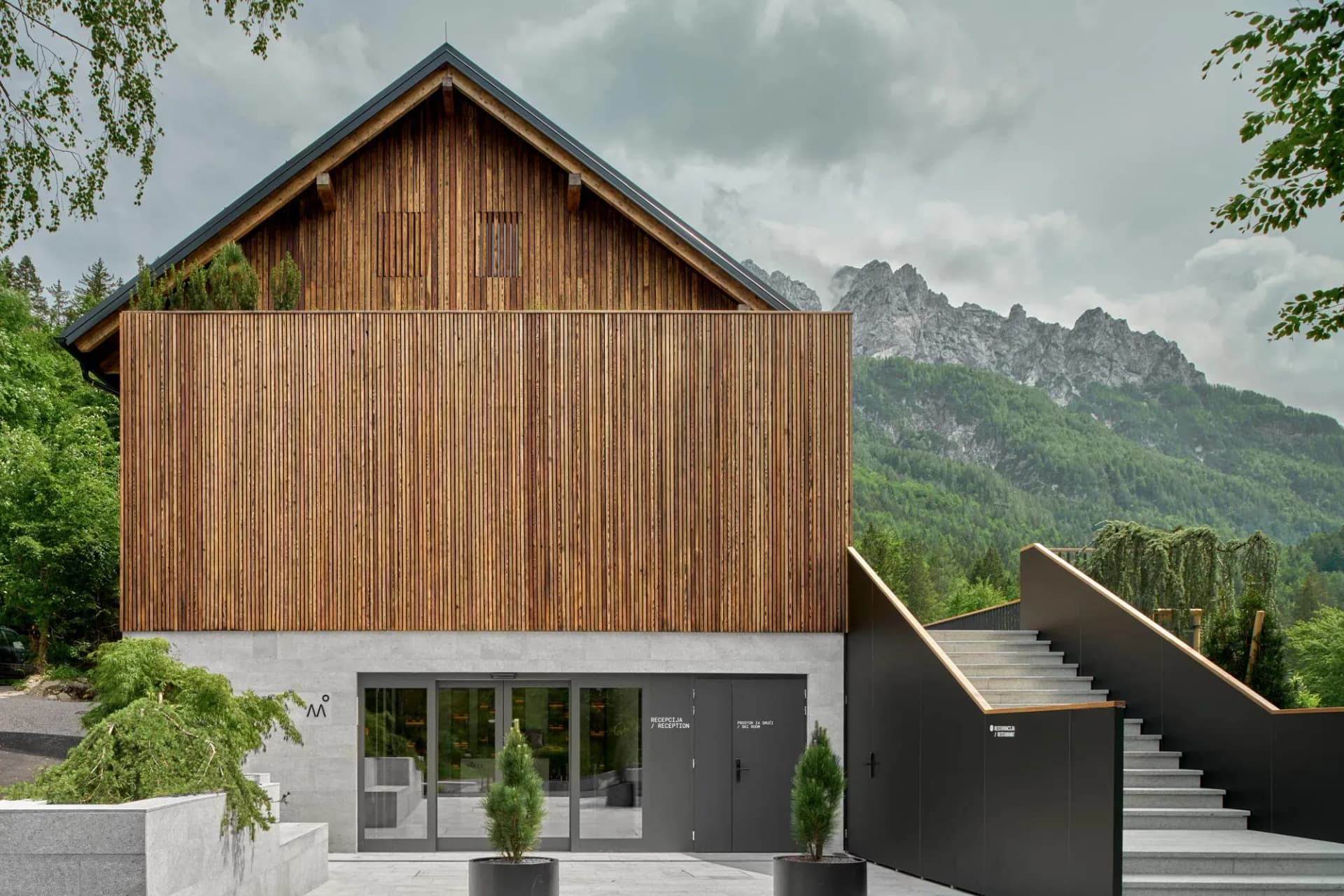 Modern wooden and concrete building entrance with stairs, set against a lush green mountain backdrop.