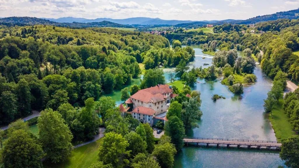 Hotel Grad Otočec aerial view, castle on river surrounded by lush green forest and hills.