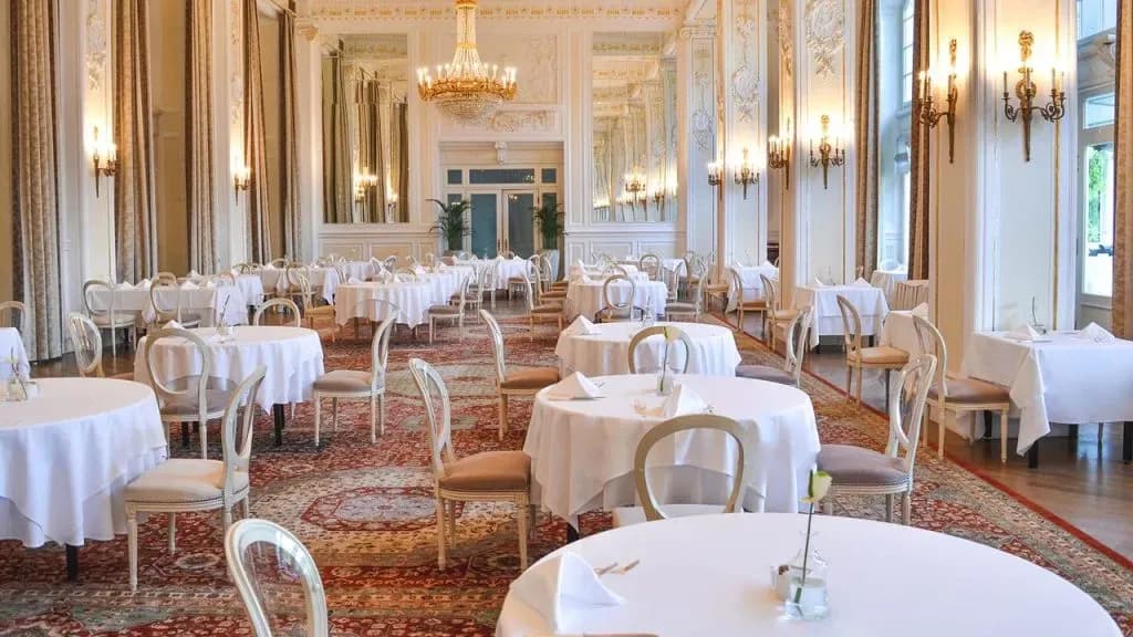Elegant dining room at Hotel Kempinski Palace set with white-linened tables and ornate chairs.