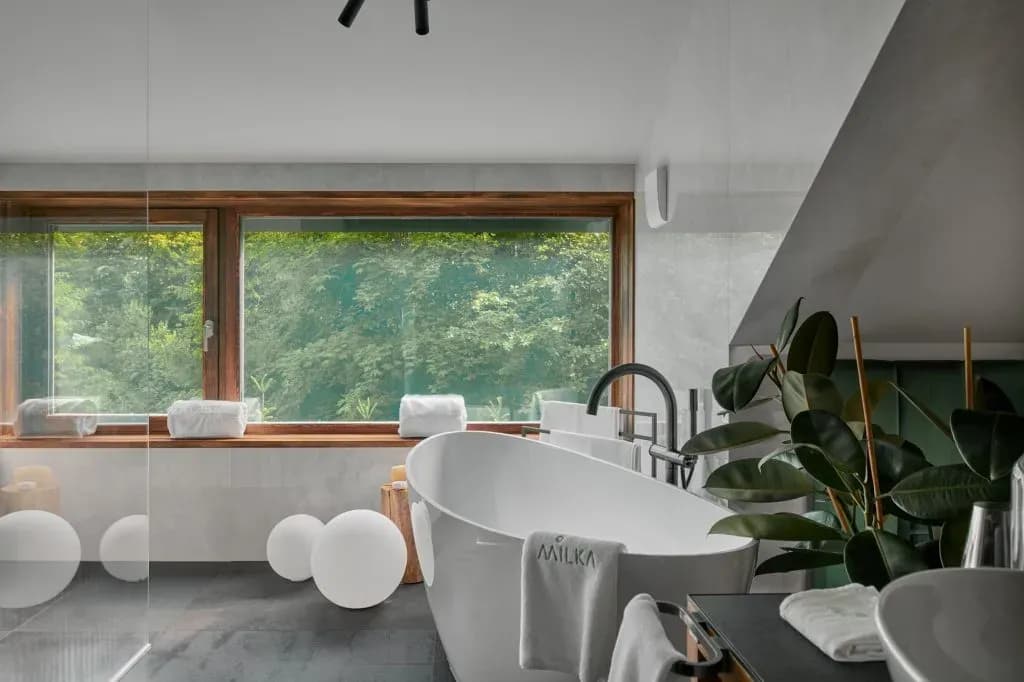 Modern bathroom with freestanding tub, large window overlooking lush green forest, and decorative orbs.