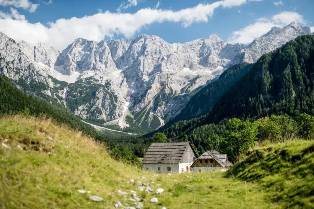 Alpine huts in green meadow below jagged, snow-dusted mountains under blue sky.