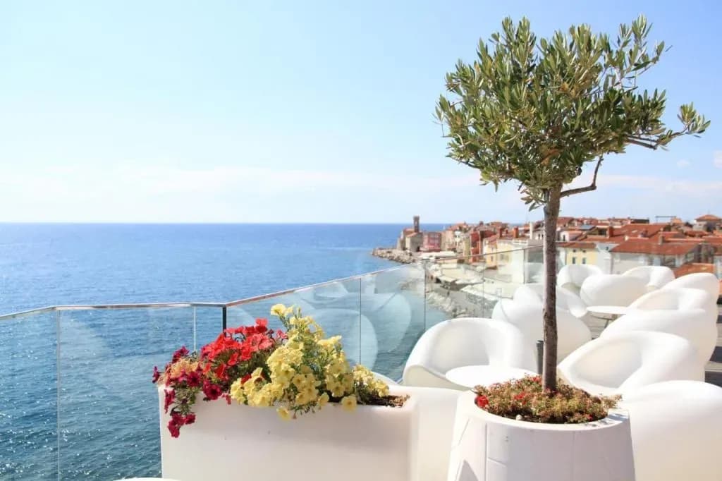 Rooftop terrace with white seating, flowers, and view of Piran coastal town and Adriatic Sea.