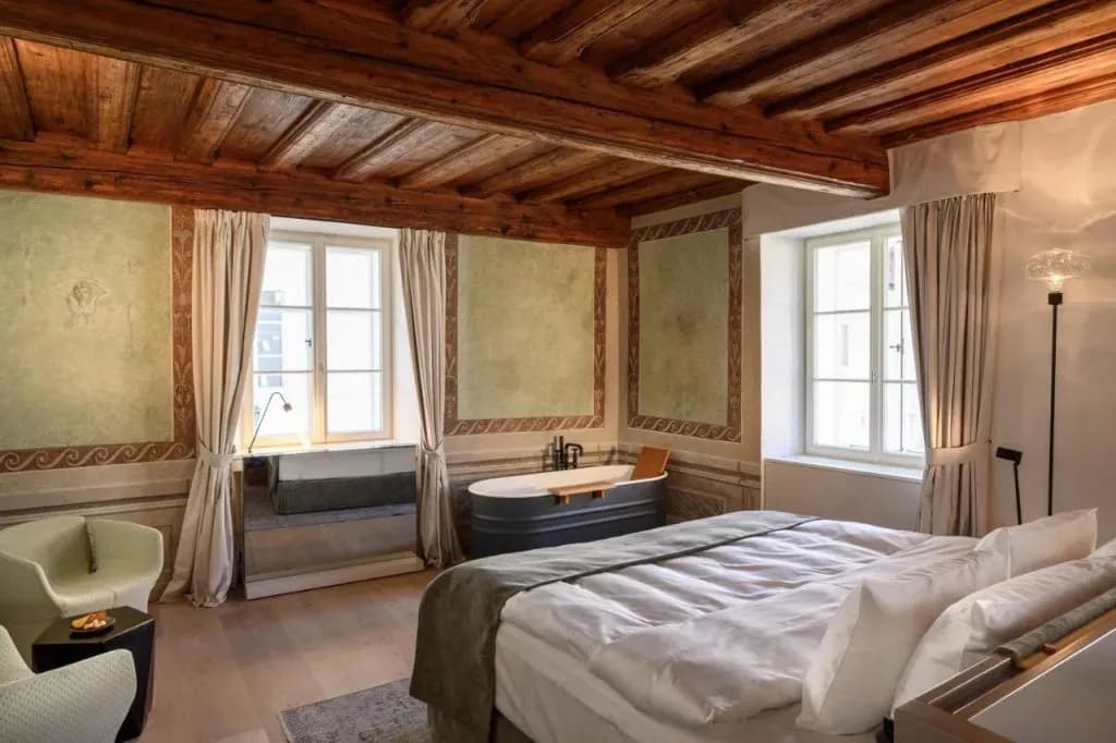 Rustic hotel room with exposed wood beams, frescoed walls, and a freestanding bathtub next to the bed.