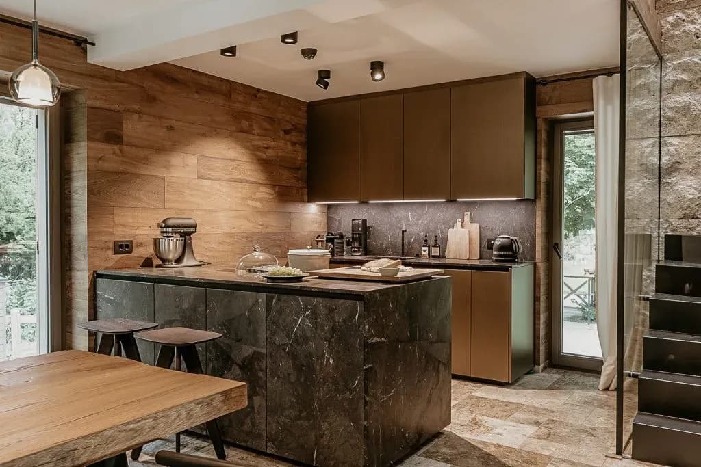 Modern kitchen with dark marble island, wood paneling, and view to greenery outside.