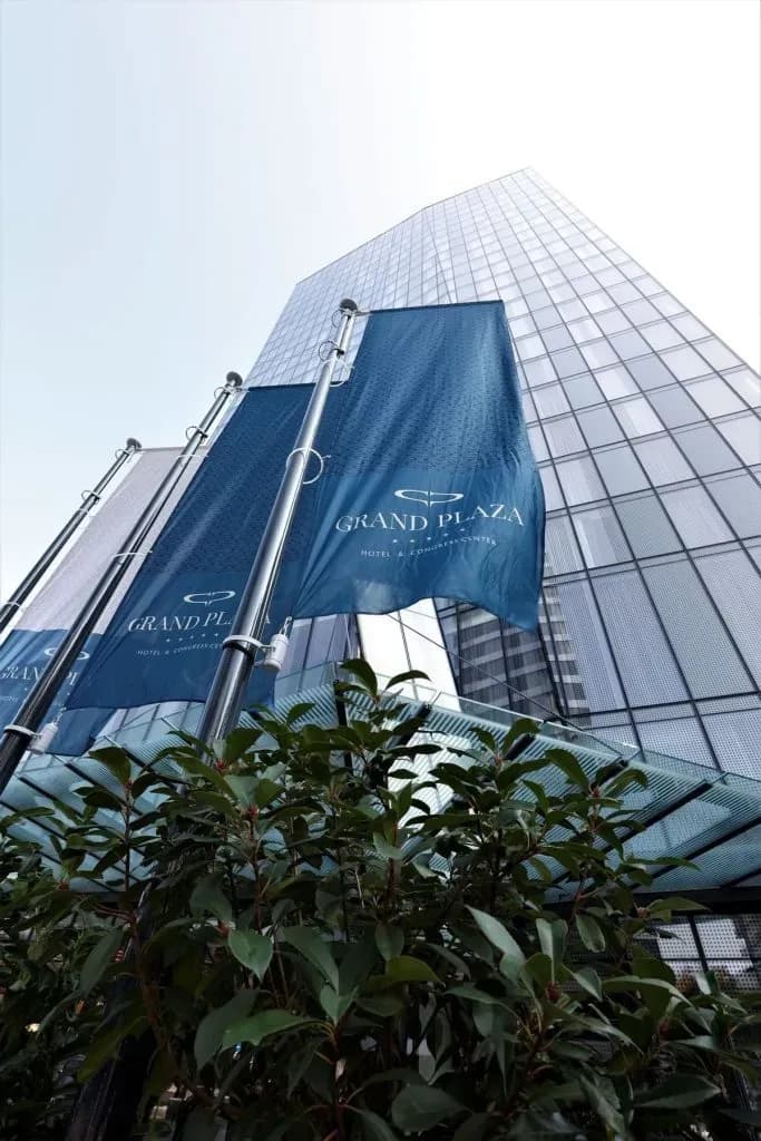 Grand Plaza Hotel flags in front of modern glass skyscraper entrance with green foliage