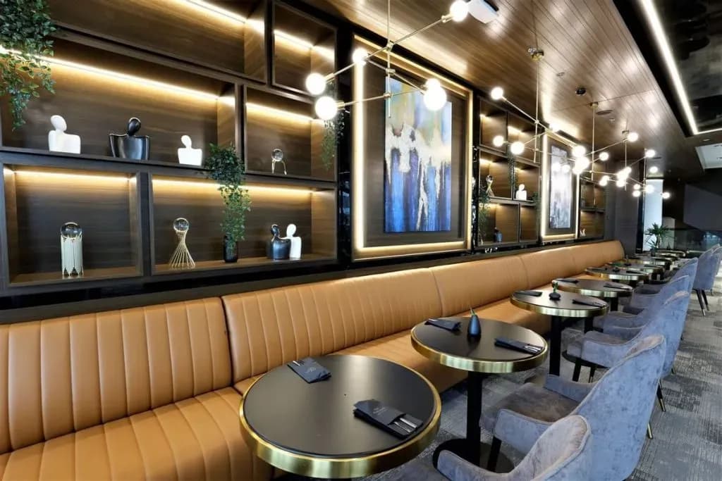 Modern restaurant interior with tan booth seating, round black tables, and illuminated shelving displaying art.