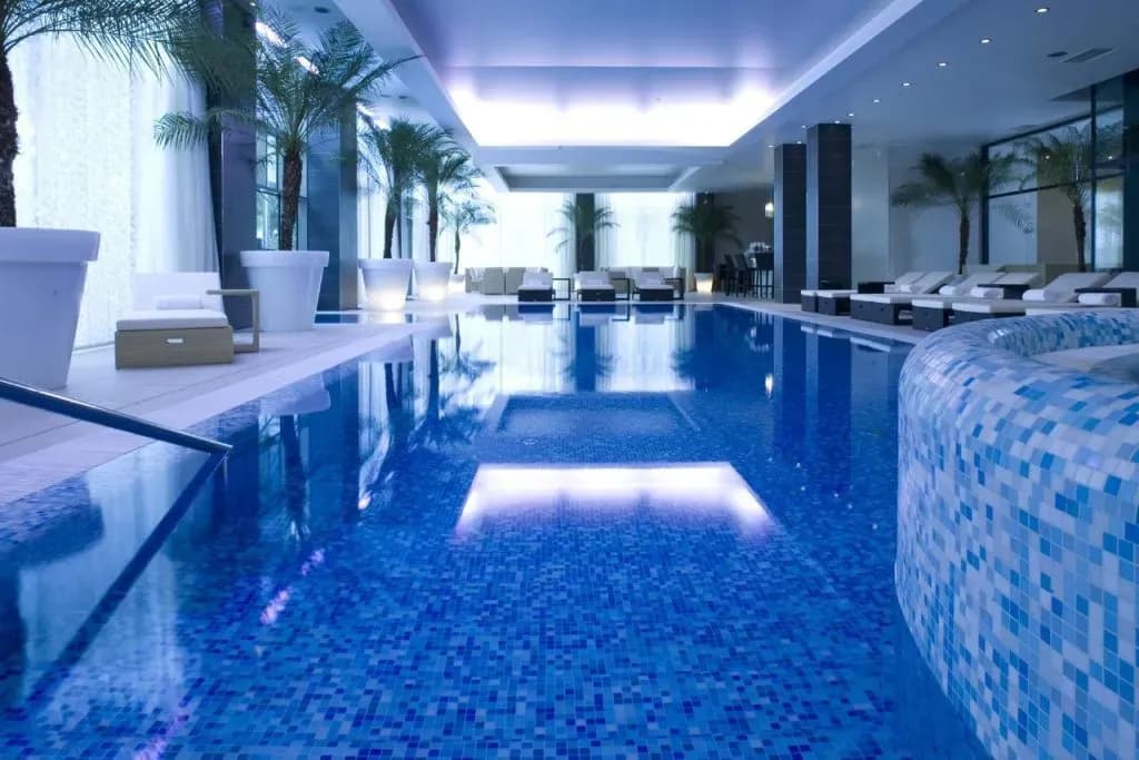 Indoor swimming pool with blue mosaic tiles, lounge chairs, and potted palm trees at Kempinski Palace.