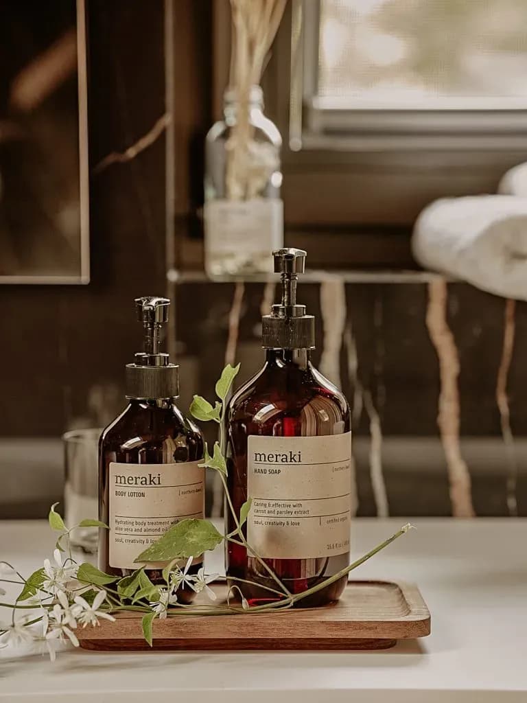 Two Meraki body lotion and hand soap bottles on a wooden tray with white flowers.