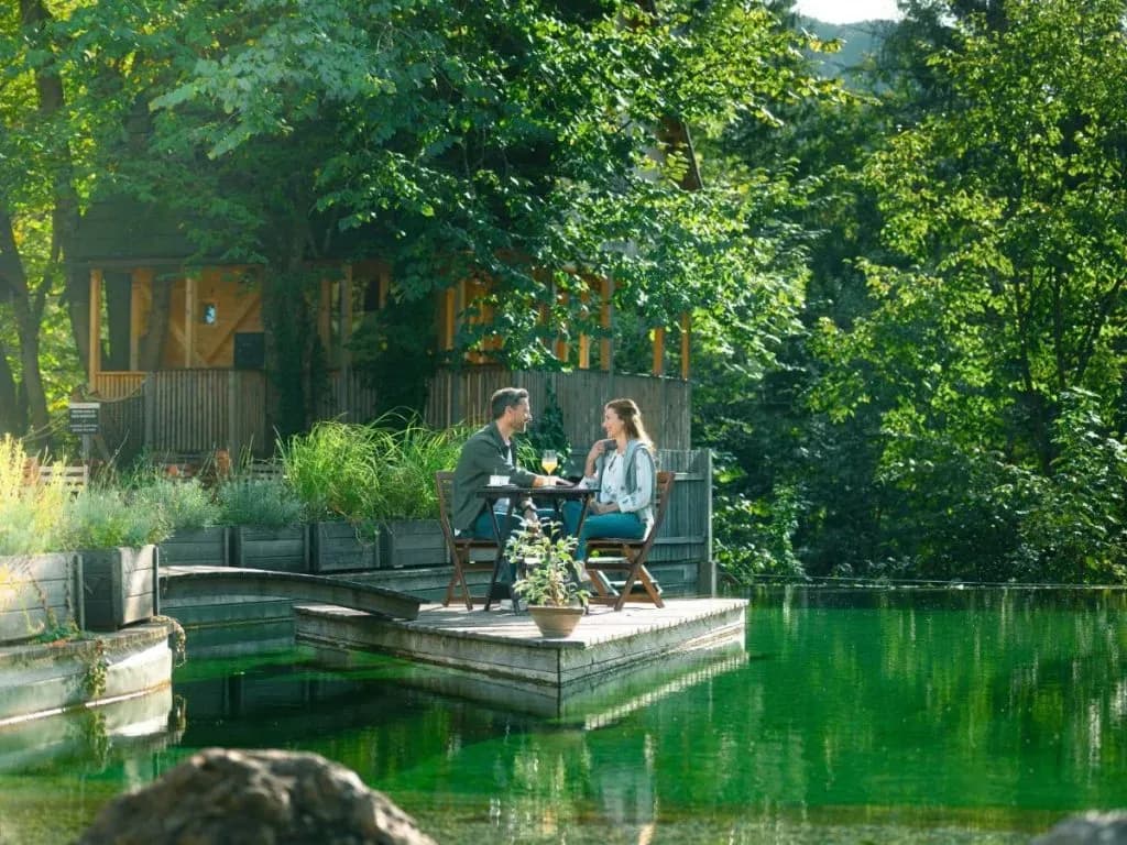 Couple dining on wooden deck over green water near wooden cabin in lush forest setting