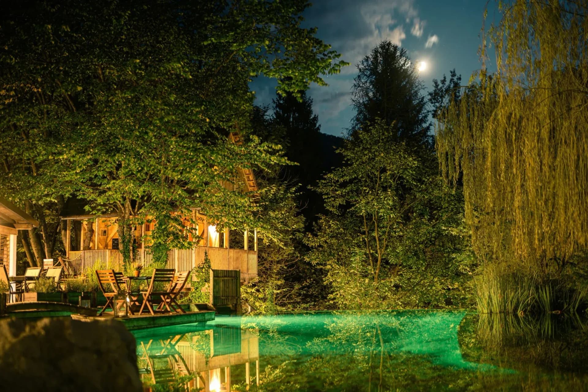 Glamping night adventure with illuminated wooden cabin by glowing green water under the moon