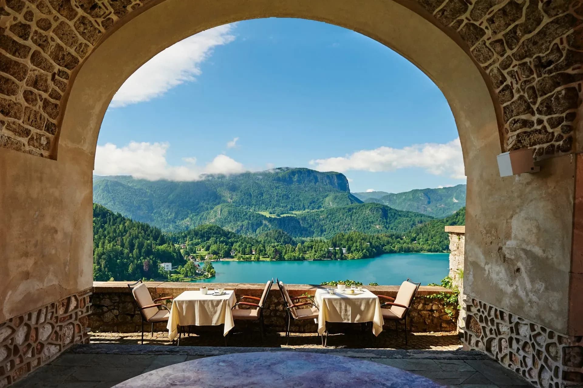 Outdoor dining tables framed by stone arch overlooking Lake Bled and forested mountains