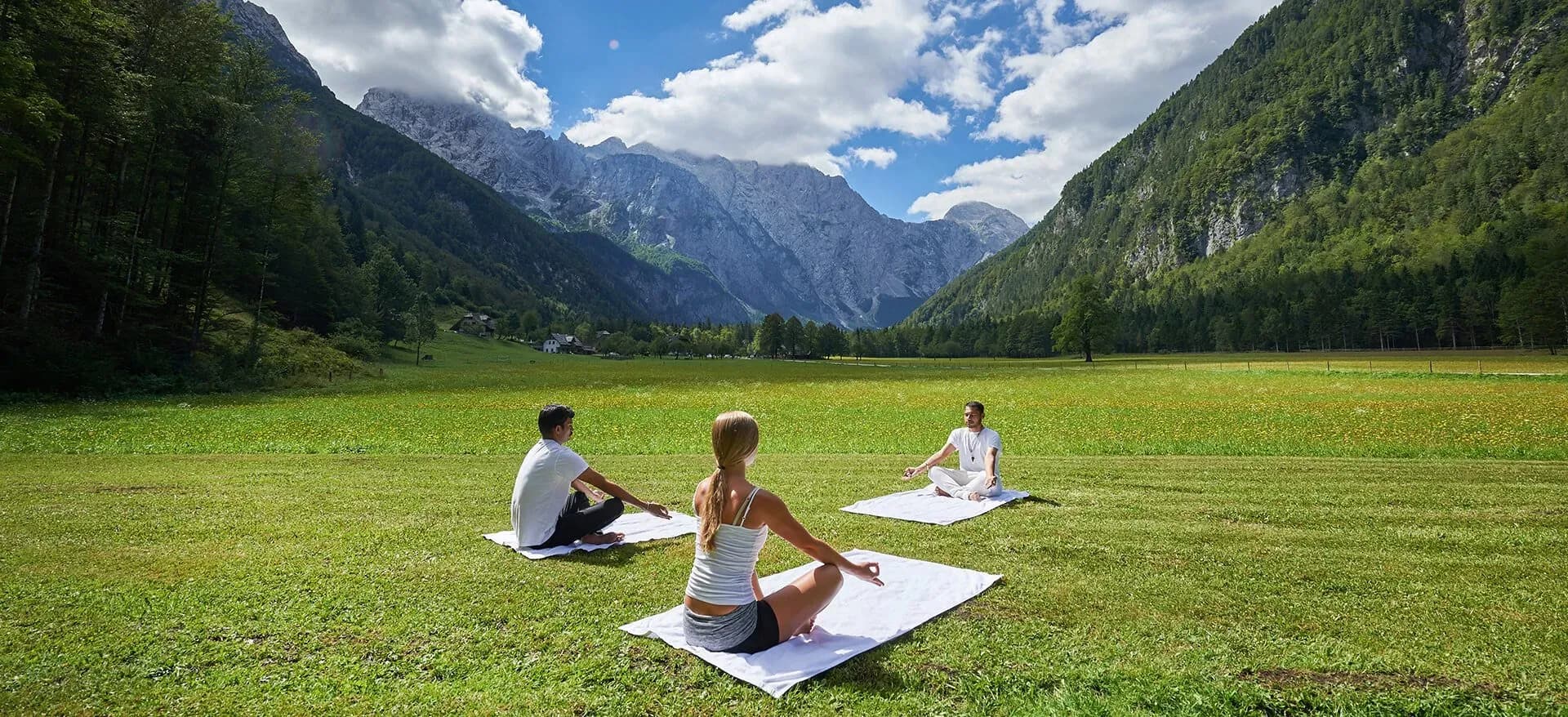 People practicing yoga outdoors in a grassy meadow surrounded by steep, forested mountains.