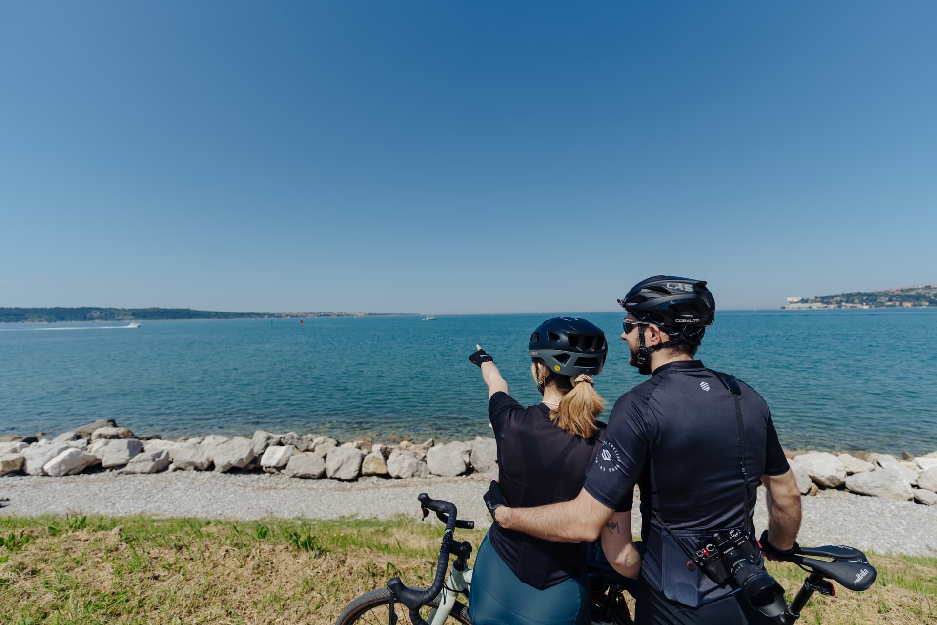 Cyclists with bikes pointing toward the sea with a distant coastline under a clear blue sky.