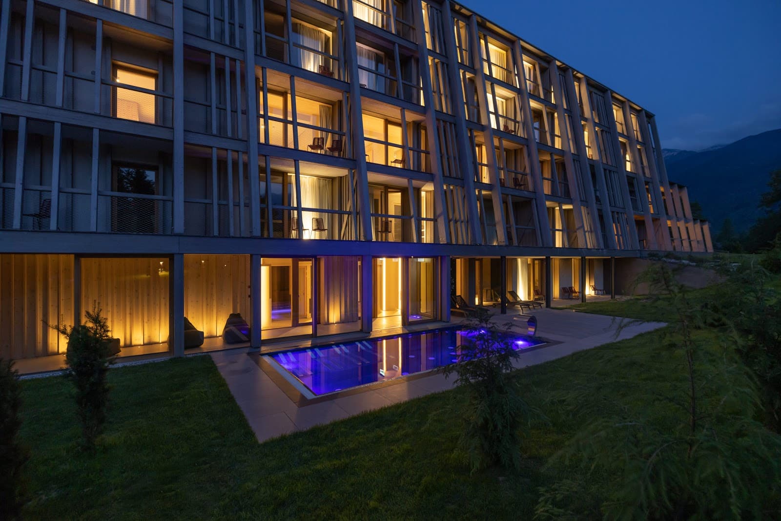 Modern hotel exterior at night with illuminated windows and a glowing blue outdoor pool near mountains.