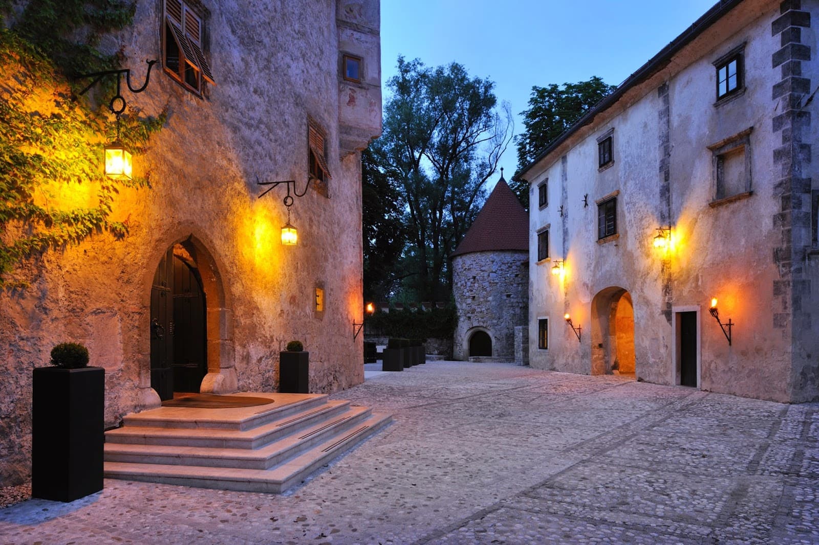 Courtyard of historic stone buildings with illuminated lanterns at dusk, Otočec.