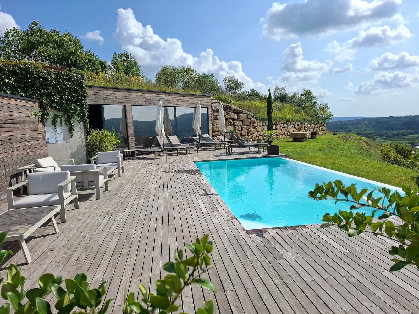 Swimming pool with wooden deck, lounge chairs, and rolling green hills under a blue sky.