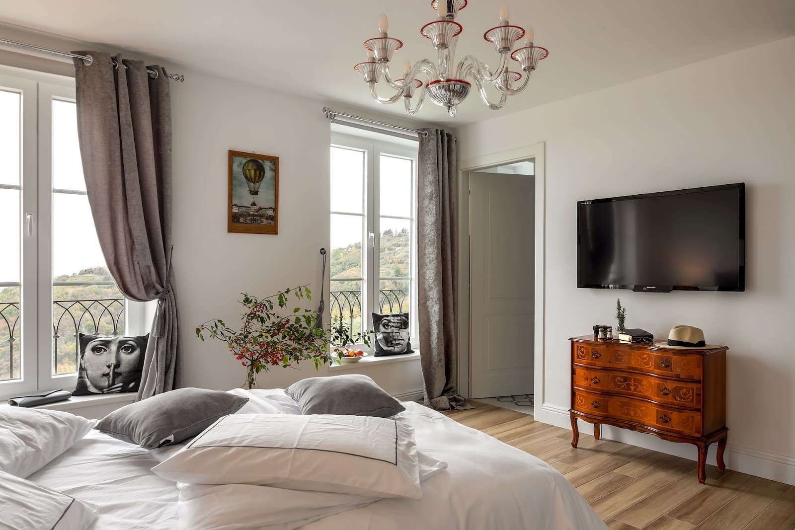 Bedroom with white linens, wood floor, chandelier, and view of rolling hills through window.