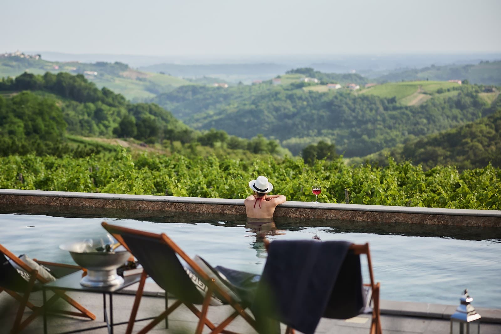 Woman in infinity pool overlooking lush green vineyard hills with glass of wine.