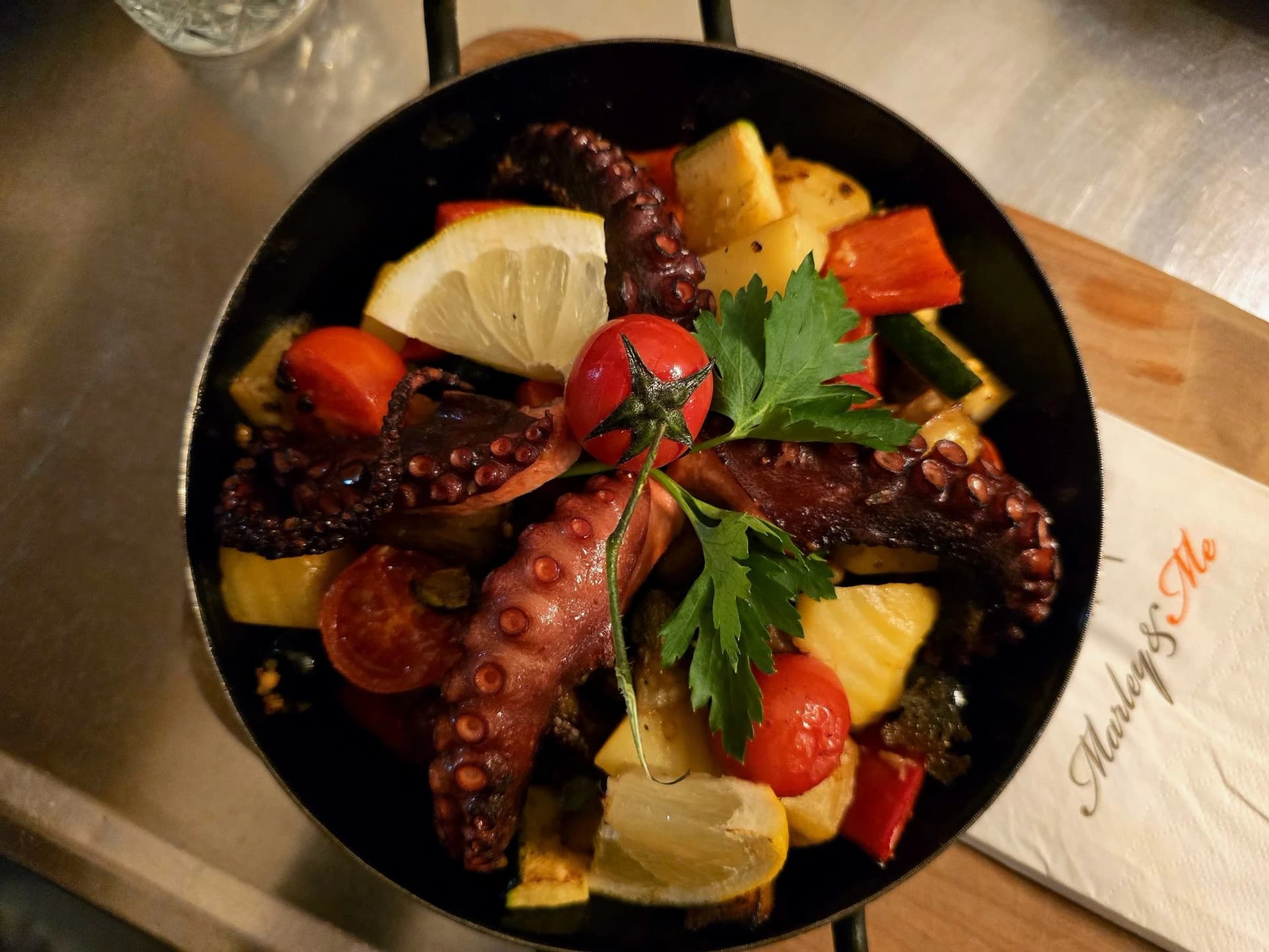 Grilled octopus with potatoes, peppers, tomato, and lemon served in a black dish next to a "Marley & Me" napkin.